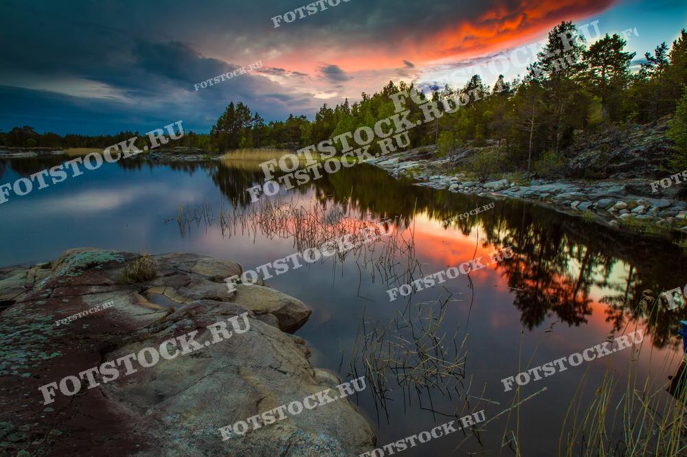 На фотографии изображен красивый закат на Ладожском озере в Карелии. Отражение закатного неба в воде, камыши и каменные берега создают атмосферу умиротворения. Вечернее небо дополняет пейзаж Ладожских шхер, подчеркивая их природную красоту.