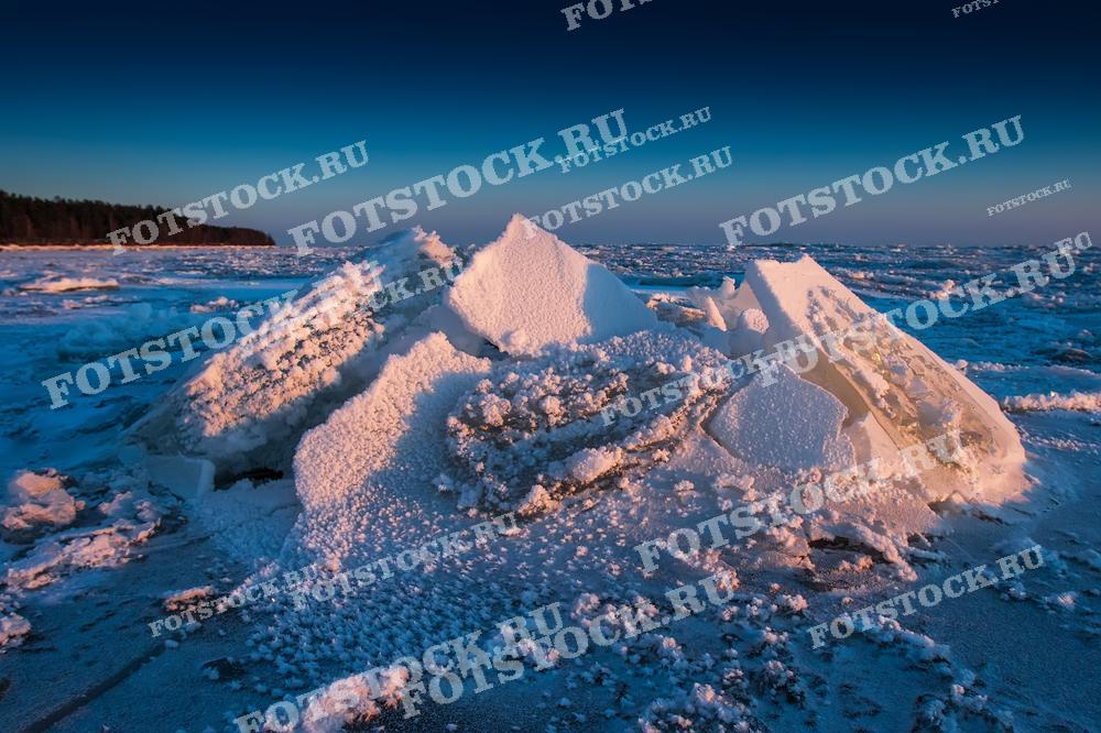 На фотографии изображен зимний берег Карелии, где массивные льдины покрыты снегом и освещены мягким светом заката. Ледяные глыбы, выдвинутые на берег, создают уникальный и впечатляющий пейзаж. Вдали виден лес, добавляющий глубину этому холодному, но живописному северному виду. Закатное солнце придает сцене теплый оттенок, контрастирующий с холодом льда.