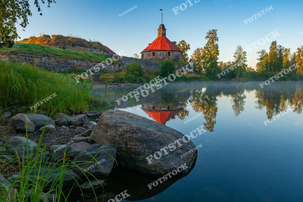 На снимке изображена крепость Корела в городе Приозерск, Республика Карелия. Это одно из древнейших фортификационных сооружений Северо-Запада России, история которого насчитывает более 700 лет. Каменные стены и башня с красной кровлей отражаются в спокойной воде, создавая атмосферу тишины и величия. Архитектурный памятник окружён зеленью и идеально вписывается в природный ландшафт карельского края.