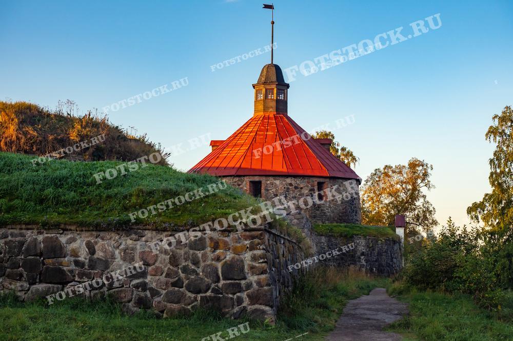 На фотографии изображена крепость Корела в Приозерске, Карелия, ранним утром. Каменные стены крепости и красная крыша башни освещены первыми лучами солнца. Крепость окружена зелеными деревьями и травой, что придает ей особое очарование. Это историческое место привлекает множество туристов благодаря своей богатой истории и живописному расположению.