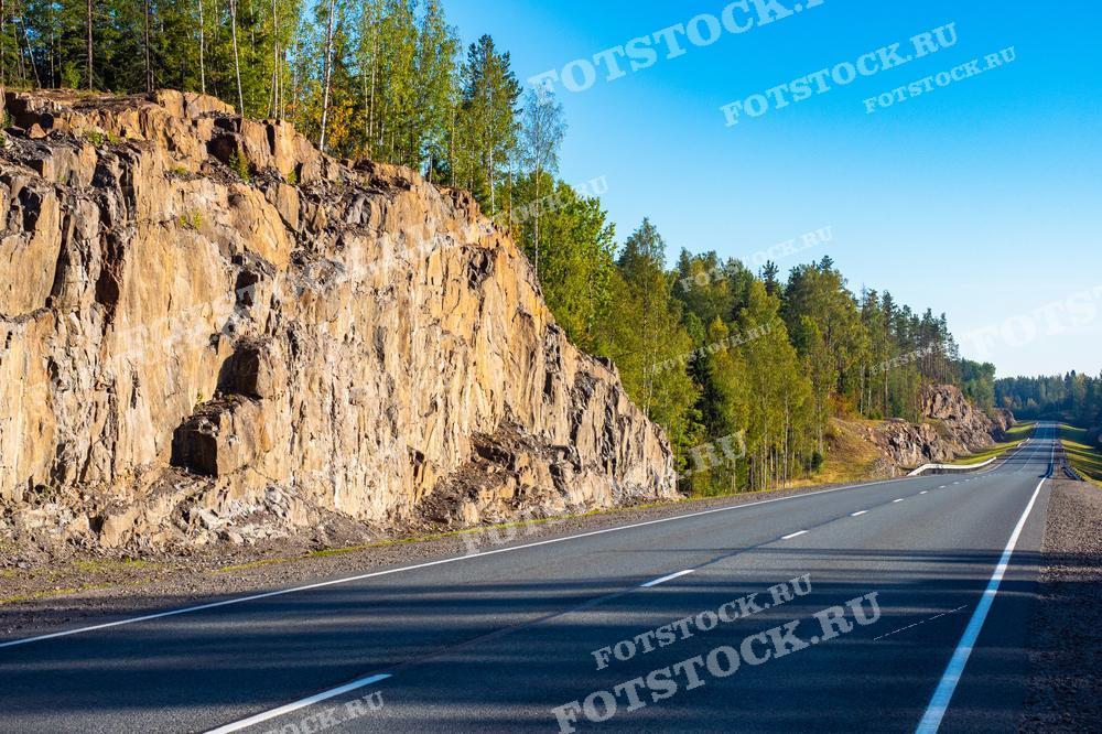 На этой фотографии изображена дорога в Карелии, проходящая мимо высоких скальных откосов. Яркий солнечный день освещает гладкий асфальт и высокие скалы. Деревья вдоль дороги добавляют зеленый контраст к серым и коричневым оттенкам скал. Такой пейзаж может быть использован в публикациях о путешествиях по России, о природе Карелии или в материалах о строительстве и инфраструктуре.