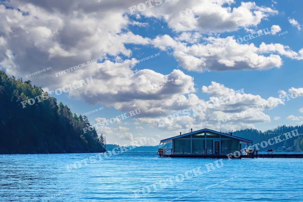 На фотографии изображён дом на воде, расположенный в Ладожских шхерах, Карелия. Плавучий дом окружён живописными лесистыми берегами и спокойными водами Ладожского озера. Облака на фоне голубого неба добавляют особую атмосферу и глубину снимку.