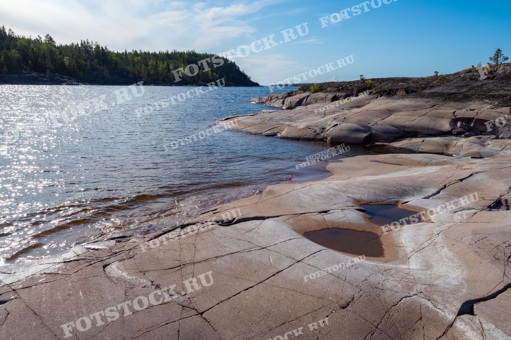 На фотографии изображены скальные шхеры Ладожского озера в Карелии, освещённые солнцем. Каменистые берега с небольшими водоёмами и лесистые участки на горизонте создают умиротворяющий пейзаж. Вода Ладожского озера блестит под солнечными лучами, придавая снимку живописный вид.