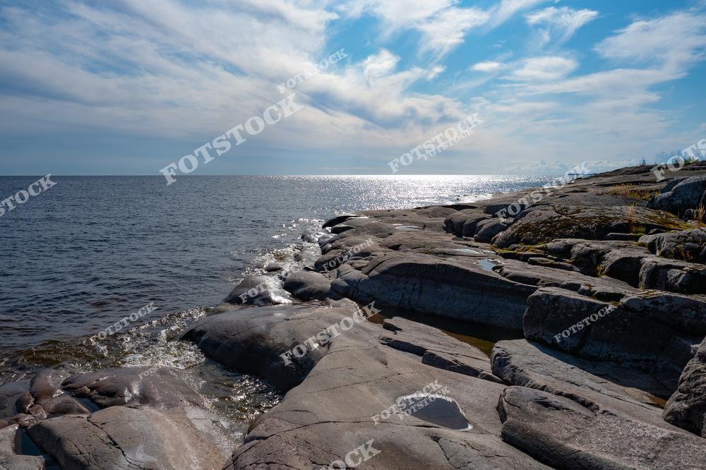 На фотографии изображён солнечный день на берегу Ладожского озера, где скалистая поверхность плавно переходит в водную гладь. Свет отражается на волнах, придавая пейзажу яркость и живость. Этот каменистый берег — типичный ландшафт Ладожских шхер, привлекающий туристов своей уникальной природной эстетикой. Тишина и просторы Ладоги делают это место идеальным для спокойного отдыха на природе.