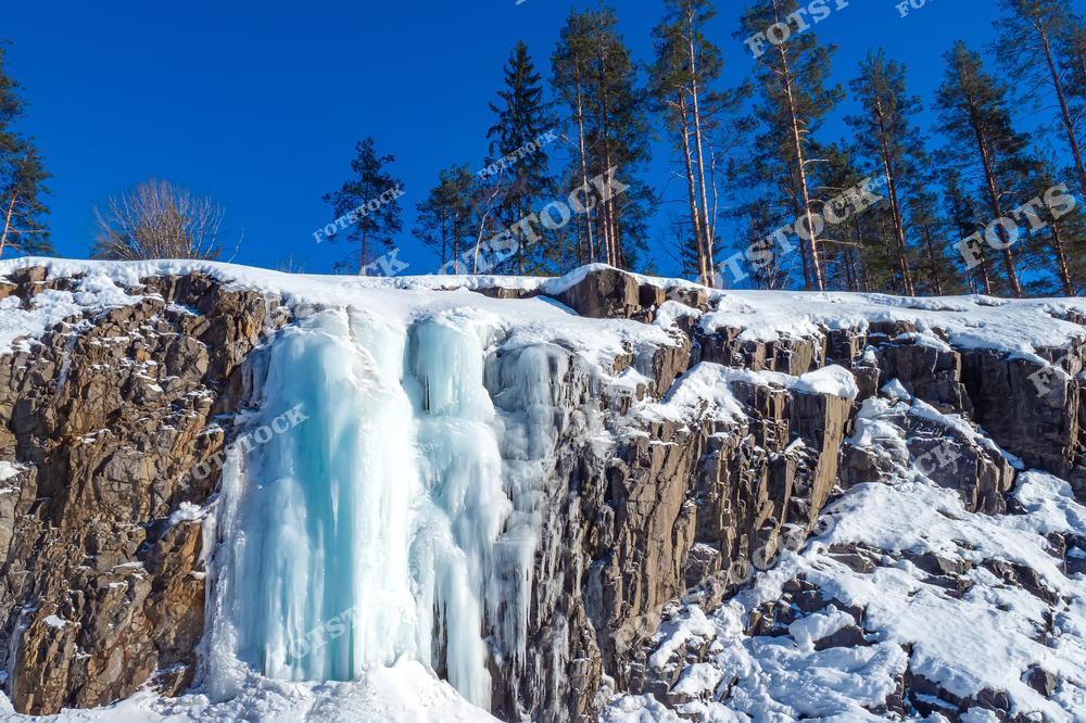 На фотографии изображён замёрзший водопад в Карелии, окружённый скалами и сосновым лесом. Ледяные массы образовали величественные каскады, сверкающие на ярком зимнем солнце. Голубое небо и белоснежный снег создают контраст, делая пейзаж особенно ярким. Такие водопады являются популярной достопримечательностью Карелии зимой, притягивая любителей природы и зимнего туризма.