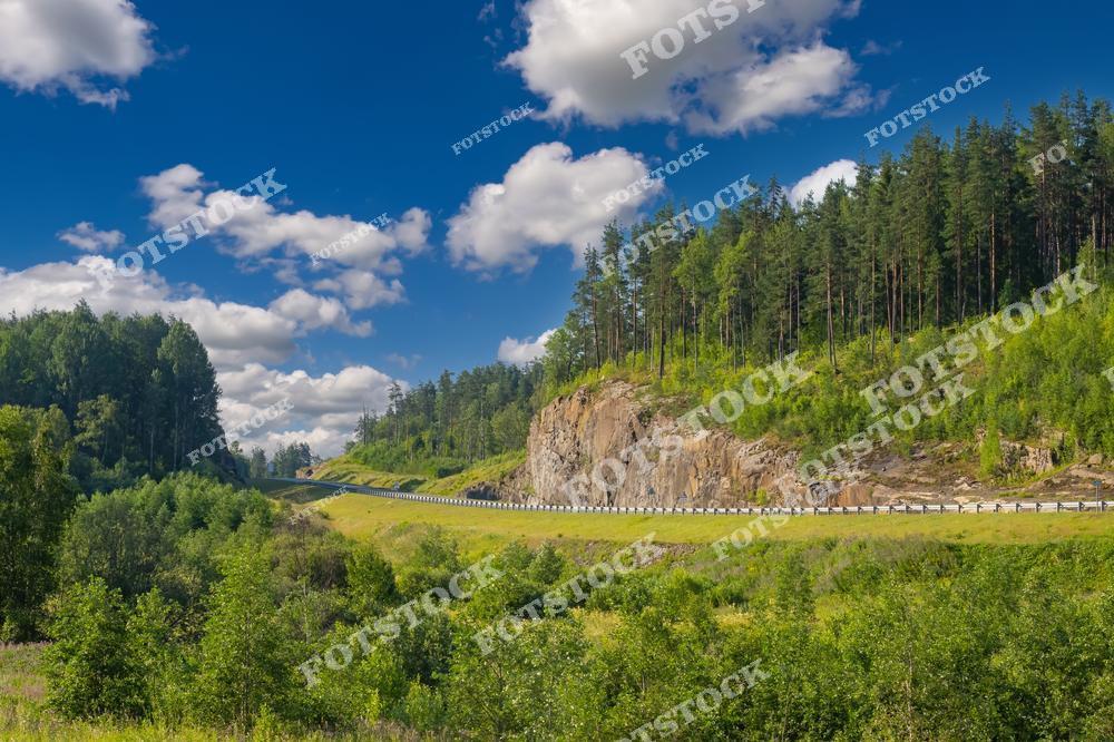 Фотография запечатлела участок автомобильной дороги в Карелии, пролегающий среди скалистых утесов и густых хвойных лесов. Голубое небо с пушистыми облаками подчеркивает летнюю атмосферу, а извилистая трасса гармонично вписывается в природный ландшафт. Это место идеально подходит для автопутешествий и знакомства с карельской природой. Многие туристы выбирают этот маршрут, направляясь к Ладожскому озеру и другим достопримечательностям региона.
