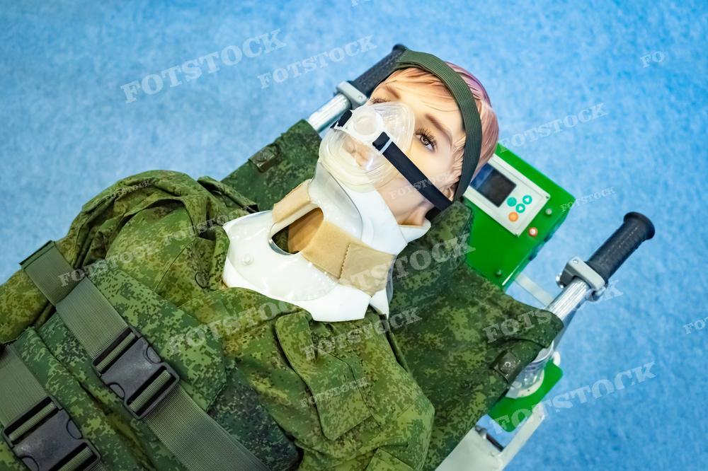 На фотографии представлен медицинский тренажёр, имитирующий пациента в маске для ИВЛ (искусственной вентиляции лёгких), лежащего на кушетке. Вокруг тренажёра размещены различные медицинские приборы и элементы военной униформы, что может указывать на тренировку военных медиков или обучение в условиях имитации полевого госпиталя. Такое фото может быть использовано в учебных целях для медицинских курсов, в статьях о военной медицине, в методических пособиях по оказанию первой помощи или интенсивной терапии.