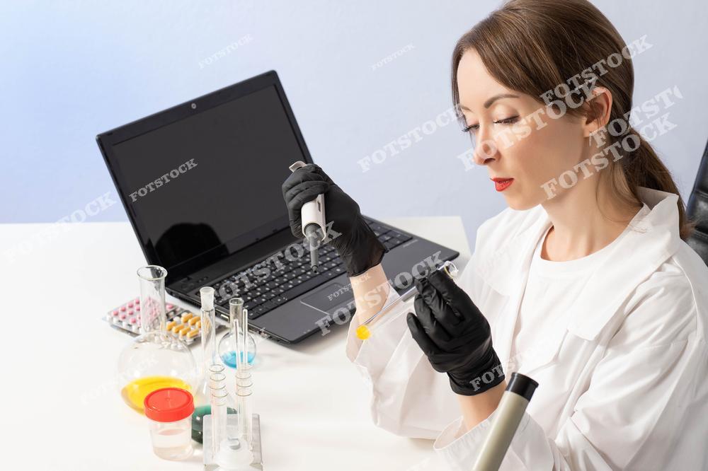 На изображении женщина в белом халате и черных перчатках работает с пробиркой и пипеткой за столом с лабораторным оборудованием. На заднем плане находится открытый ноутбук, символизирующий использование технологий в исследовании. Фото подчеркивает внимание к деталям и профессиональный подход к лабораторным экспериментам.