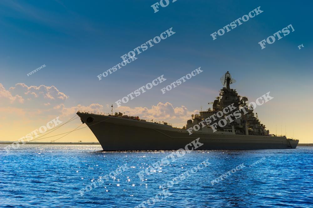 На фотографии изображён военный корабль, стоящий на воде под лучами закатного солнца. Силуэт корабля подчёркивает его внушительные размеры и мощь на фоне тёплого неба и спокойного моря. Снимок иллюстрирует красоту и значимость военно-морского флота.
