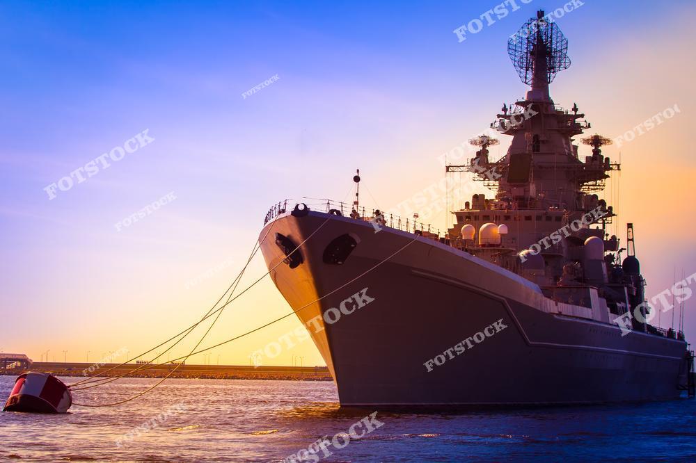 На фотографии изображён крупный военный корабль, пришвартованный на воде в лучах закатного солнца. Тёплые оттенки закатного неба подчёркивают силуэт судна, символизирующего мощь и технологичность военно-морского флота. Снимок демонстрирует красоту и значимость современных морских технологий.