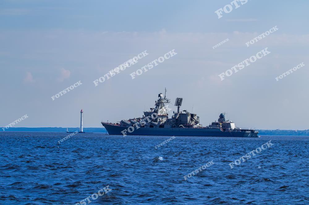 На изображении военный корабль, стоящий в спокойных водах моря, с маяком на заднем плане. Светлое голубое небо и горизонт создают атмосферу простора и безопасности. Снимок символизирует морскую мощь, службу и защиту границ.