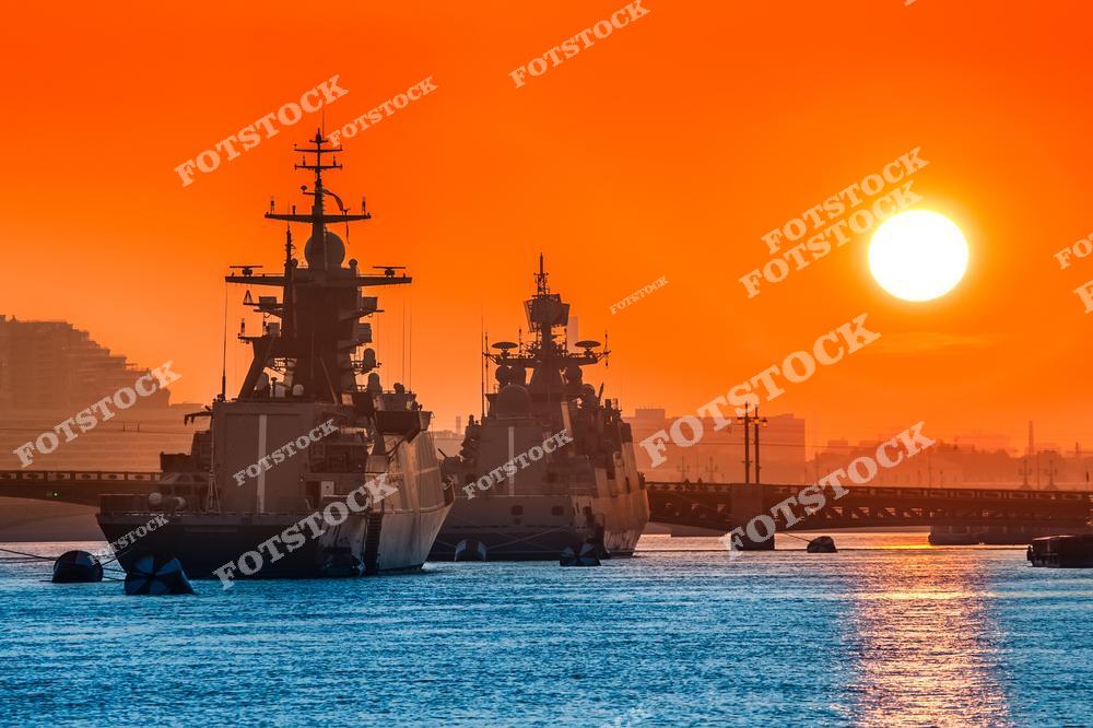На фотографии изображены два военных корабля, пришвартованных в порту на фоне яркого закатного солнца. Оранжево-красное небо подчёркивает силуэты судов, создавая драматическую атмосферу. Снимок отражает современность и стратегическую значимость военно-морских технологий.