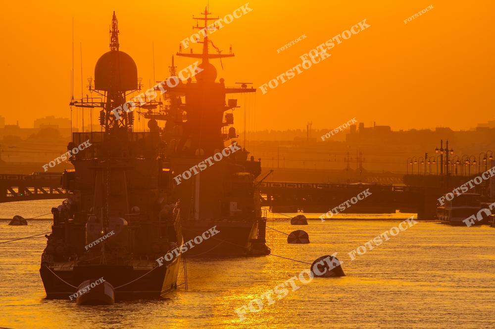 На фотографии изображены военные корабли, пришвартованные в порту на фоне золотого закатного неба. Тёплый свет солнца создаёт мягкий контраст между силуэтами кораблей и спокойной поверхностью воды. Снимок подчёркивает величие военно-морской техники в гармонии с природой.