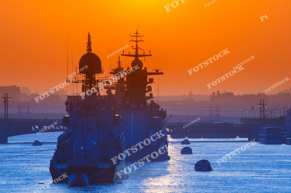 Фотография показывает пришвартованные военные корабли в порту, освещённые закатным солнцем. Глубокий оранжевый цвет неба подчёркивает силуэты кораблей и отражается в спокойной воде. Снимок демонстрирует баланс между инженерной мощью и природной красотой.
