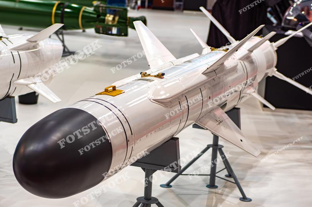 На изображении представлена современная тактическая ракета, установленная на стенде выставки военной техники. Экспонат демонстрирует новейшие достижения в области вооружений. На заднем плане видны другие образцы техники и оружия.