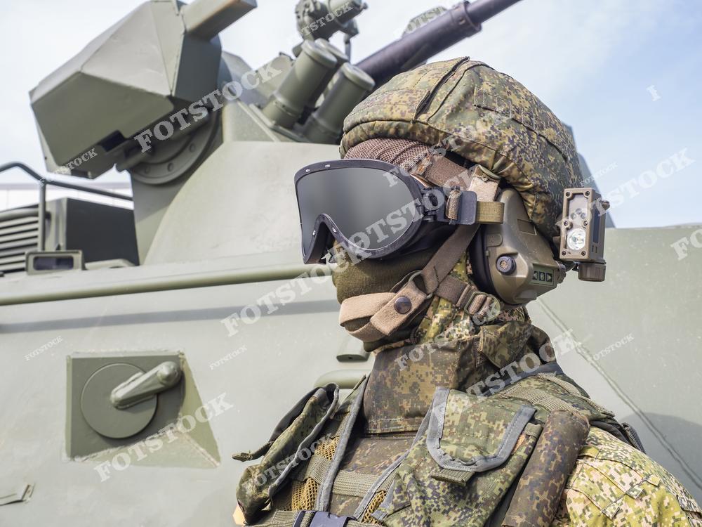 На фотографии изображен военный в камуфляжной форме, защитных очках и шлеме, стоящий рядом с бронетехникой. Оснащение подчеркивает уровень подготовки и техническую оснащенность вооруженных сил. Снимок символизирует современные технологии и тактическую готовность.