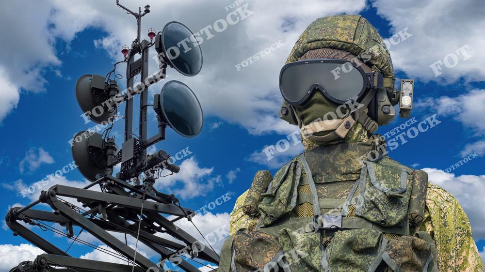 На фотографии изображен военный в камуфляже и защитной экипировке, стоящий рядом с высокотехнологичными антеннами на фоне неба. Изображение символизирует современные системы связи и тактическое оснащение вооруженных сил. Кадр передает атмосферу готовности и технической продвинутости.