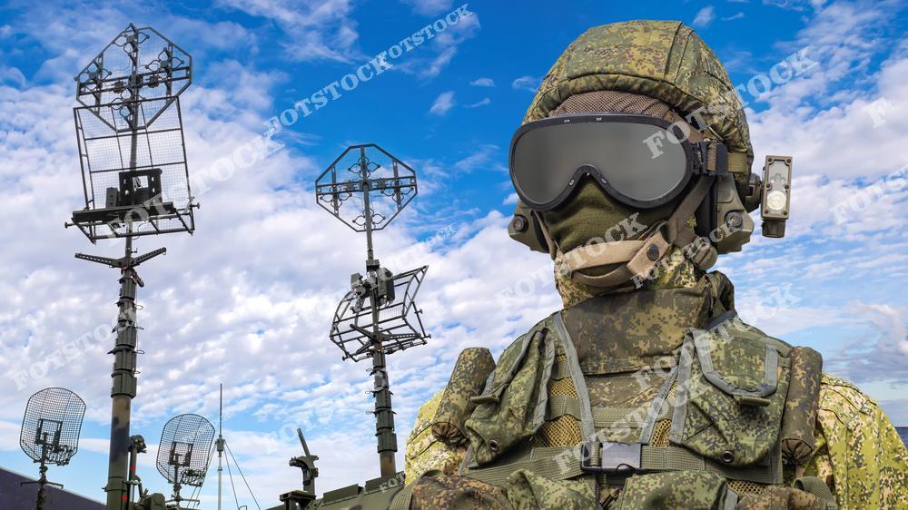 На изображении представлен солдат в современном военном обмундировании с закрытым лицом и защитными очками. За ним находятся радарные установки и элементы ракетной системы на фоне голубого неба. Фотография символизирует современные технологии и обороноспособность ракетных войск.