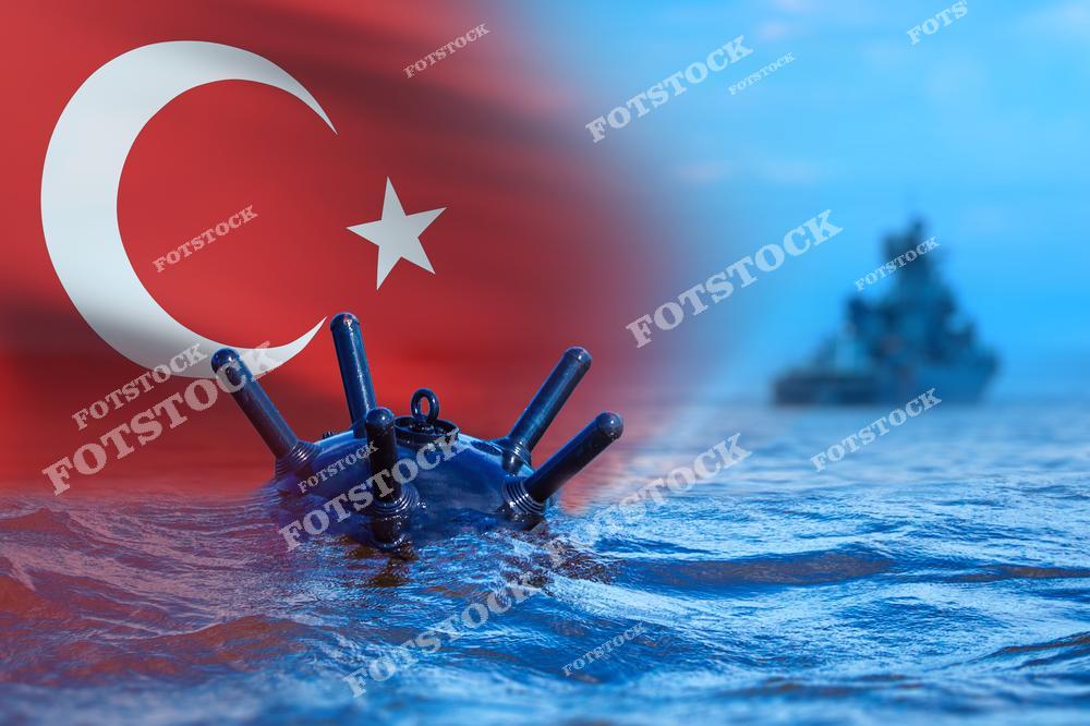 На переднем плане изображена морская мина, находящаяся на поверхности воды, с флагом Турции в левой части изображения. Вдалеке виден военный корабль, патрулирующий территориальные воды. Снимок символизирует морскую безопасность, оборону и стратегическую важность контроля морских границ.