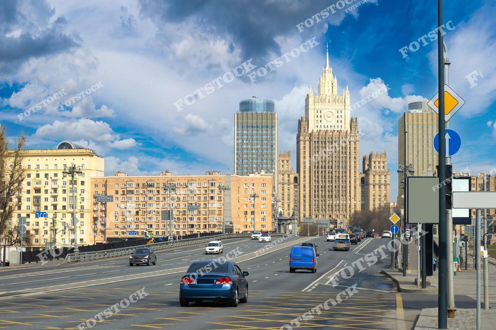 На фото изображена одна из центральных улиц Москвы с магистралью, по которой движутся автомобили. Вдалеке видна сталинская высотка — Министерство иностранных дел России, дом, выполненный в характерном для советской эпохи архитектурном стиле. Высокие современные здания, окружающие старинный памятник архитектуры, подчёркивают контраст между исторической и современной архитектурой Москвы. Яркое небо и облака создают живописный фон для динамичной городской сцены.