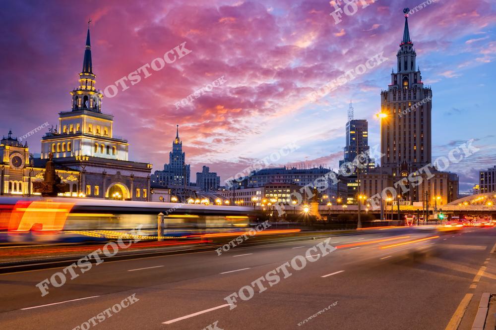 На фотографии запечатлён вечерний момент в Москве, когда закат окрашивает небо в яркие цвета. В центре внимания — оживлённая улица, на которой машины создают эффект движения. Это изображение хорошо передаёт атмосферу московской жизни, где исторические здания сочетаются с динамичным движением транспорта. Вдали можно увидеть московскую архитектуру, включая здания в стиле сталинского ампира и современные высотки. Такая панорама отражает контраст между историей и современностью в столице России.