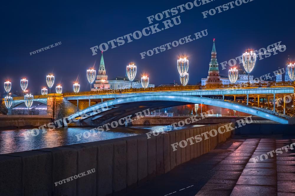 На фотографии изображён зимний вечерний вид на Москву. Мост через Москву-реку украшен праздничными гирляндами в форме огоньков, создающих атмосферу торжества и праздника. Вдалеке видны Кремль и красиво подсвеченные исторические башни. Такое изображение может быть использовано для статей о праздновании Нового года и Рождества в России, в туристических брошюрах и как декоративный элемент в оформлении интерьеров, подчёркивающий красоту московских зим.