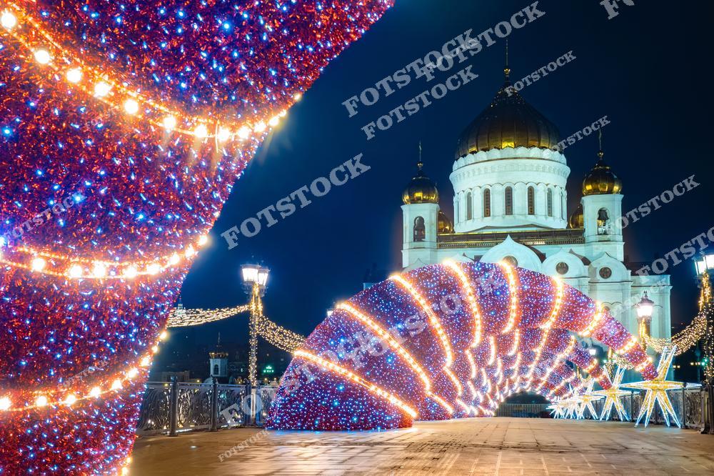 На фотографии изображён Храм Христа Спасителя в Москве, который в новогоднюю ночь украшен яркой иллюминацией. Окружающие улицы украшены светящимися арками и звездами, создающими волшебную атмосферу праздника. Праздничные огни отражаются в ночном небе, придавая дополнительное сияние этому историческому и культурному символу столицы. Храм Христа Спасителя, один из самых значимых православных храмов в России, в этом контексте выглядит ещё более величественно и празднично.