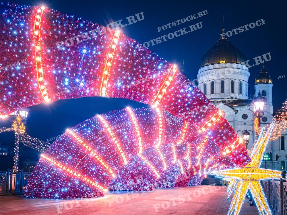 На фотографии изображены яркие рождественские огни и украшения на фоне Храма Христа Спасителя в Москве. Это фото может быть использовано для иллюстрации праздничного оформления города, в рекламных материалах, посвящённых Рождеству и Новому году, а также для туристических путеводителей.