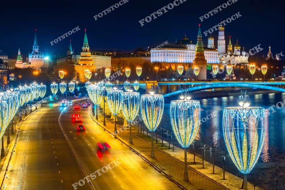 На фото изображена Москва в ночь, когда город украшен праздничной иллюминацией. В центре композиции — Кремль, освещённый огнями, с яркими башнями и историческими зданиями, создающими величественную атмосферу. Вдоль реки Москва расположены новогодние украшения в виде светящихся элементов, создающих эффект волшебства. Вдалеке виден мост, соединяющий две части города. Улицы оживлены движущимися автомобилями, что добавляет динамики в этот зимний вечер.