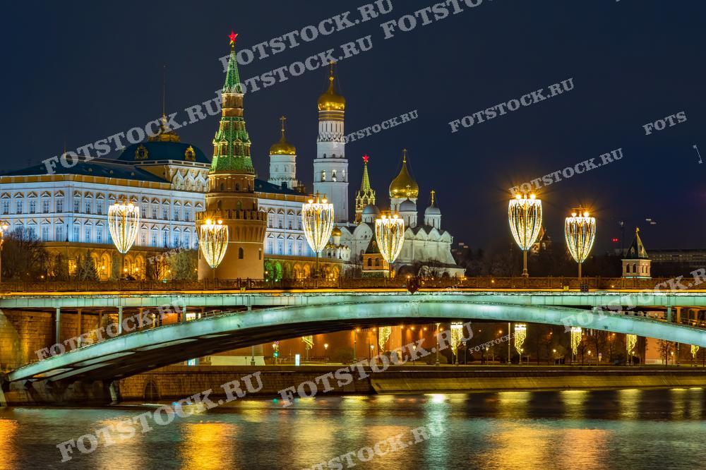 На фотографии запечатлен ночной вид на Москву с яркими украшениями моста и подсветкой Кремля во время новогодних праздников. Отражение праздничных огней в тёмных водах реки придаёт сцене волшебное ощущение. Это изображение может быть использовано для оформления статей о новогодних и рождественских традициях в России, для туристических проспектов, предлагающих зимние путешествия в Москву, или как часть декоративного оформления в кафе и ресторанах, создающих атмосферу праздника.