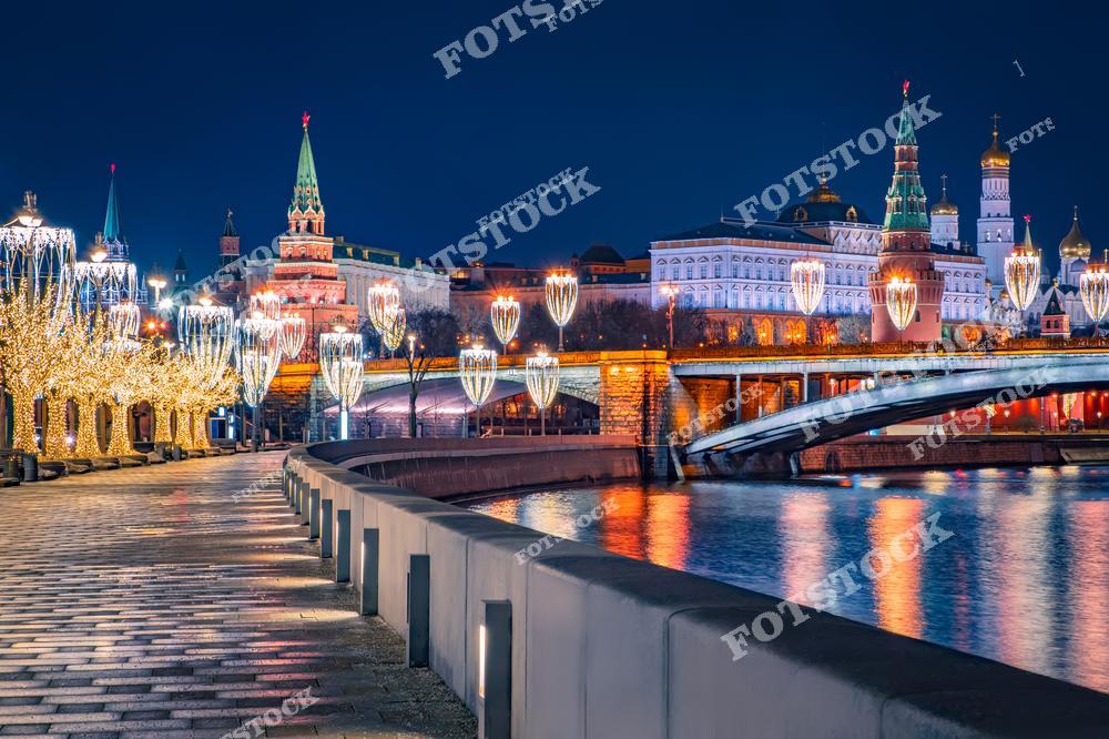 На фото изображён вечерний вид на Московский Кремль, украшенный яркой новогодней иллюминацией. Вдоль набережной реки Москва стоят светящиеся ёлки и декорации, создающие атмосферу праздника. Мост, соединяющий два берега реки, также украшен огнями, отражающимися в воде. Кремлёвские башни, включая Спасскую, подсвечены яркими огнями, что подчеркивает величие и историческую значимость этого архитектурного комплекса. Закатный свет и праздничная иллюминация делают атмосферу особенно волшебной.