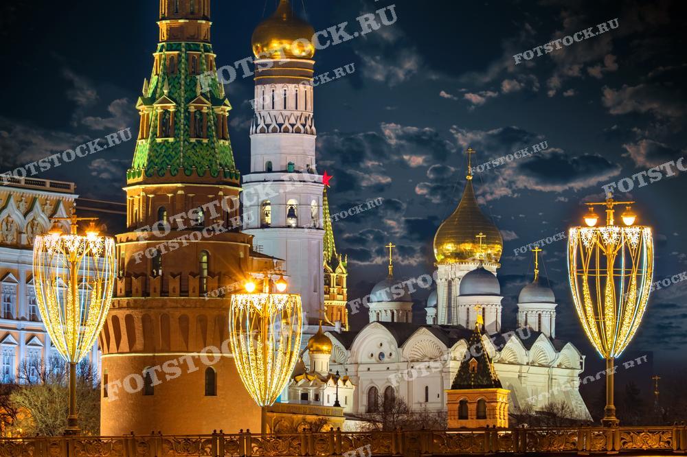 На фотографии запечатлен зимний вечер в Москве с видом на кремлёвские башни и собор. Величественные здания Кремля освещены и подсвечены, что подчёркивает их архитектурное великолепие и историческое значение. Праздничные огни на переднем плане добавляют атмосферу торжества и праздника, делая изображение идеальным для использования в материалах, связанных с рождественскими и новогодними праздниками в России. Фотография может быть использована в туристических брошюрах, культурных публикациях и как элемент декора.