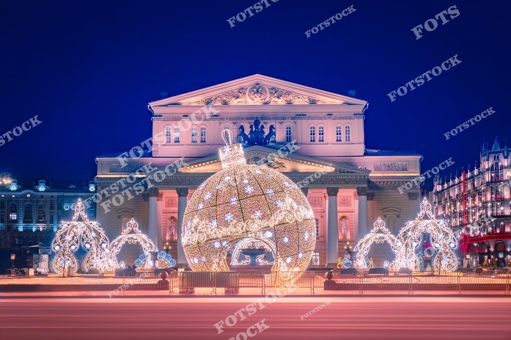 На фотографии изображен Большой театр в Москве, окруженный праздничной новогодней иллюминацией. Центральное место занимает гигантский светящийся шар и арки, украшенные гирляндами. Театр выделяется своим классическим архитектурным фасадом, гармонично сочетающимся с праздничным оформлением площади. Вечерняя подсветка и насыщенный синий фон усиливают атмосферу зимнего праздника. Композиция прекрасно передает волшебный дух новогодней Москвы.