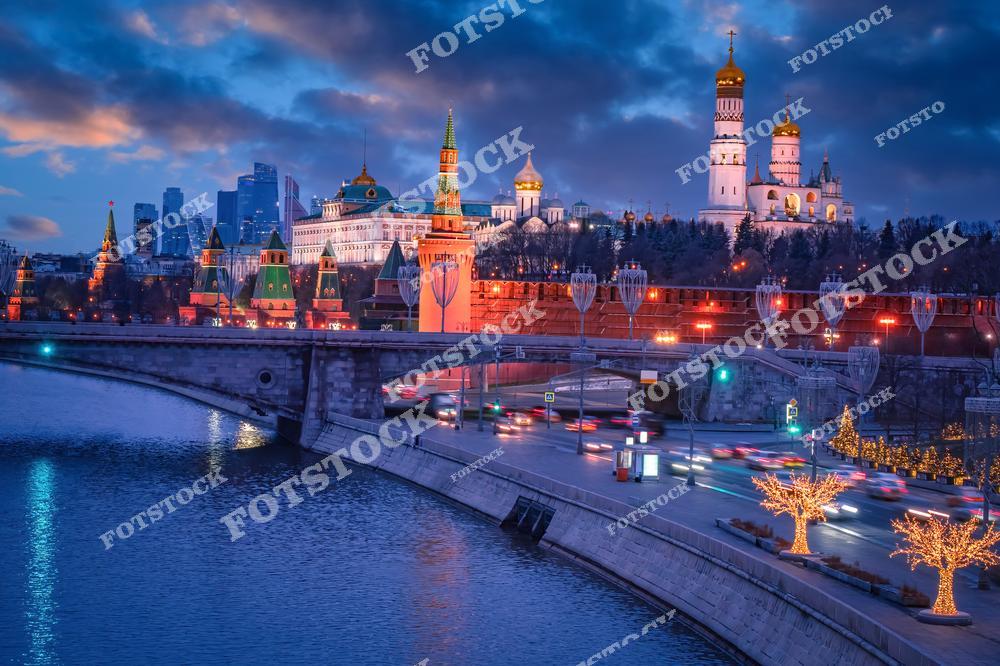 На фотографии изображён Кремль в вечернее время, освещённый праздничной иллюминацией. Хорошо видны Спасская башня, Благовещенский собор и колокольня Ивана Великого. В правой части снимка — украшенные огнями деревья вдоль набережной, создающие новогоднее настроение. Вдали виднеются современные небоскрёбы, создавая контраст с древней архитектурой Кремля. Слева проходит река Москва, отражающая огни города. На переднем плане — оживлённая дорога и мост с движущимися автомобилями.