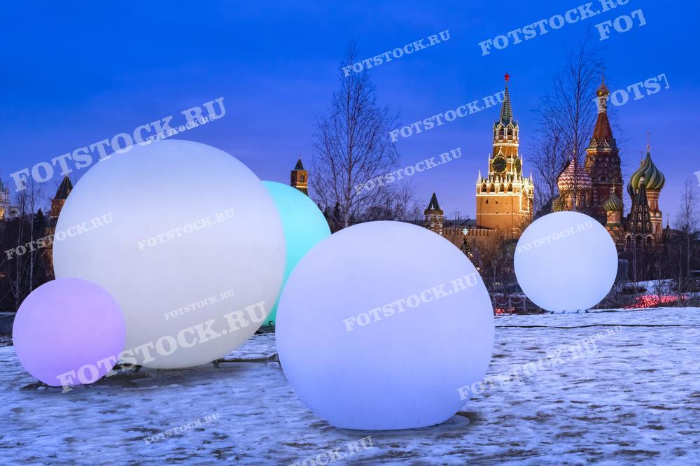 На фотографии изображены огромные декоративные светящиеся шары на фоне Красной площади в Москве в сумеречное время. Эти шары, изменяющие цвет, создают контраст с историческими кремлёвскими стенами и знаменитым Собором Василия Блаженного. Изображение может быть использовано для иллюстрации уникальных современных арт-объектов в традиционных городских условиях, в статьях о городском искусстве и культурных мероприятиях в Москве, а также в рекламных и промо-материалах для туристических агентств, подчеркивающих зимнюю красоту города.