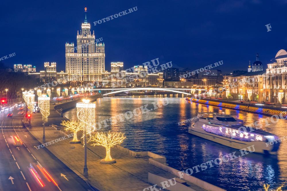 На фотографии изображена вечерняя Москва с праздничной иллюминацией. На заднем плане видна одна из сталинских высоток, освещенная огнями. Вдоль набережной движется прогулочный катер, а вдоль дороги видны украшенные деревья и светящиеся фонари.