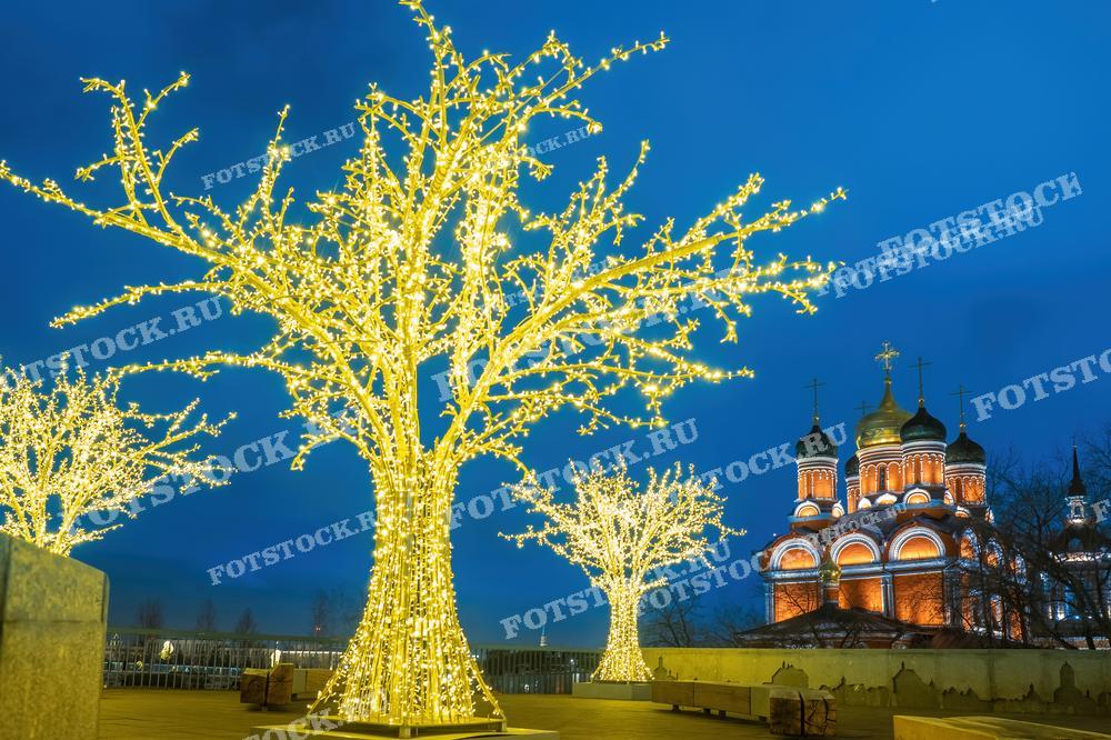 На фотографии изображены искусственные деревья, покрытые множеством светодиодных лампочек, создающие эффект заснеженных деревьев под ярким зимним небом. Эти светящиеся деревья расположены на фоне здания с исторической архитектурой, которое освещено тёплым светом, подчеркивающим его красивые купола и орнаменты. Фотография может использоваться для привлечения посетителей на зимние фестивали или мероприятия, а также в качестве иллюстрации в статьях о зимних городских праздниках.