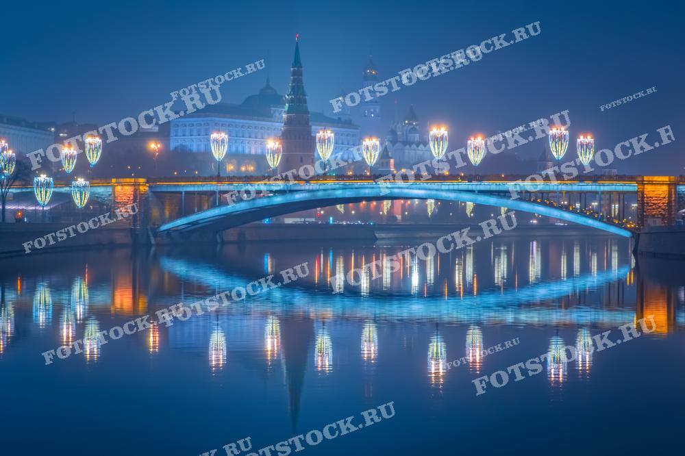 На фотографии изображен вечерний вид на Москву-реку и Большой Каменный мост, украшенный праздничной иллюминацией. Вдали виднеется Московский Кремль с подсвеченными башнями и куполами. Огни мостов и Кремля отражаются в спокойной воде реки, создавая живописный городской пейзаж.