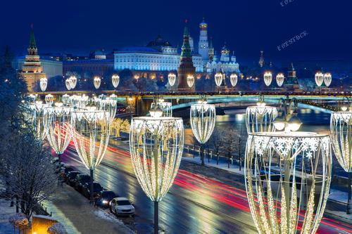На этом снимке представлено праздничное вечернее время в Москве с потоком автомобилей на Кремлевской набережной. В фоне виднеется Московский Кремль, подсвеченный вечерним небом. Фонари украшены светящимися гирляндами, создавая атмосферу новогоднего и рождественского волшебства. Эта фотография иллюстрирует уникальное сочетание архитектурной красоты, городского движения и праздничной атмосферы в столице России. Она может использоваться для иллюстрации статей о праздновании нового года в Москве, а также в рекламных материалах, связанных с туризмом и городскими событиями.