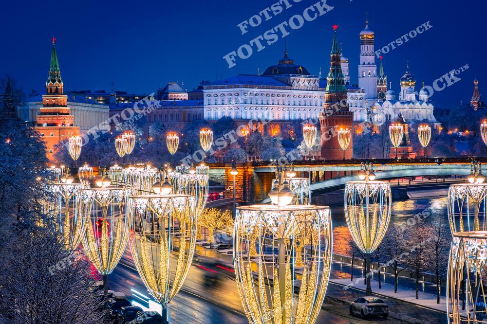 На фотографии запечатлён ночной вид на Московский Кремль, украшенный новогодней иллюминацией. Река Москва, отражающая огни гирлянд, подчёркивает красоту праздничного оформления. Кремлёвские башни и соборы, такие как Архангельский собор и колокольня Ивана Великого, создают атмосферу исторического величия. Большой Каменный мост украшен световыми конструкциями, придавая композиции волшебный зимний облик. Снежные деревья и сверкающие огни подчёркивают новогоднее настроение Москвы.