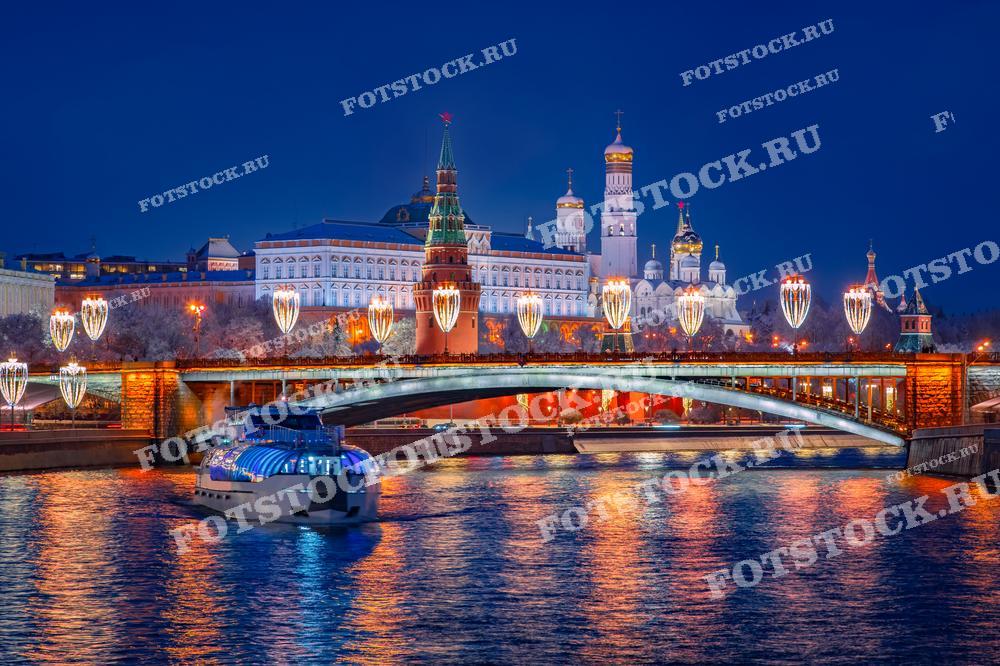 На фотографии изображен Кремль ночью, освещенный праздничной иллюминацией. В центре кадра — Большой Кремлевский дворец и колокольни Ивановской площади. Спасская башня возвышается рядом с Кремлевской стеной. На переднем плане виден один из московских мостов, украшенный новогодними гирляндами. По Москве-реке идет прогулочный катер с яркой подсветкой, создавая атмосферу праздника и зимней сказки.