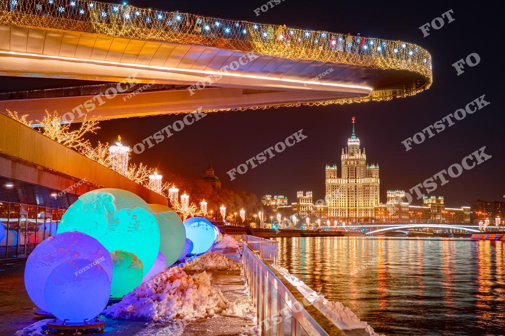 На фотографии изображён парк Зарядье с его Парящим мостом, украшенным праздничной иллюминацией. На переднем плане яркие светящиеся шары добавляют новогоднего настроения. Вдали видна сталинская высотка на Котельнической набережной, эффектно подсвеченная в ночное время. Москва-река отражает огни, придавая сцене дополнительную глубину. Это сочетание современного дизайна парка и исторической архитектуры создаёт впечатляющий городской пейзаж.