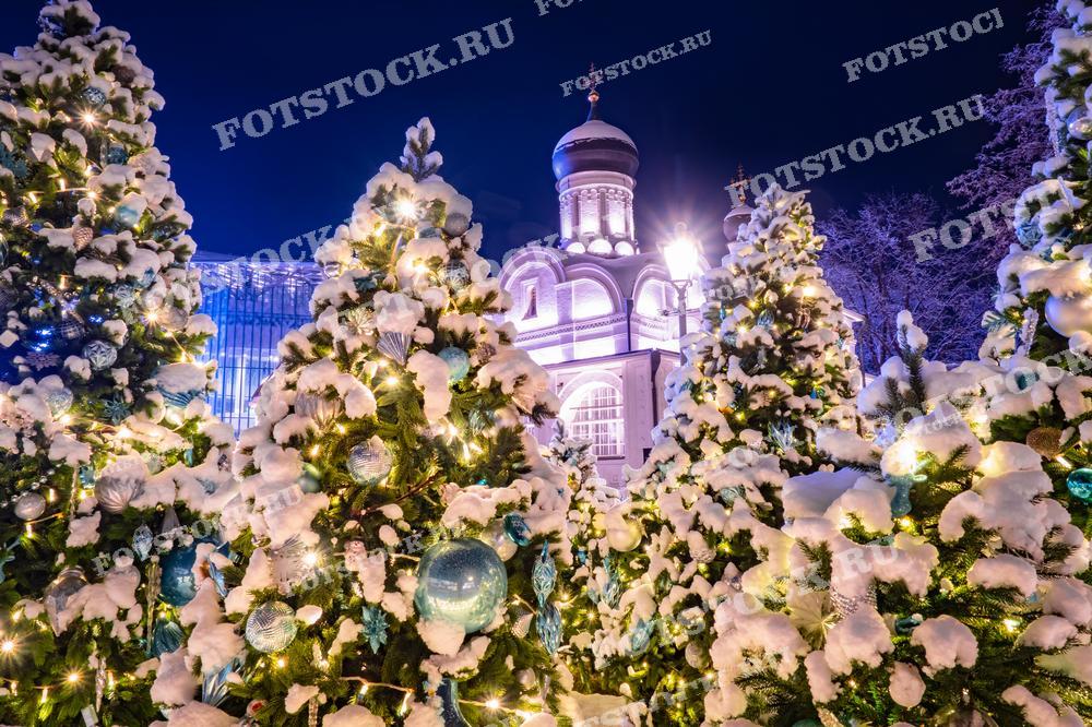 На фотографии изображены заснеженные новогодние елки, украшенные гирляндами и игрушками. На заднем плане виден храм с освещенными стенами, что добавляет торжественности и праздничного настроения. Елки усыпаны снегом и мерцают под светом гирлянд, создавая ощущение рождественского чуда. Эта сцена олицетворяет зимнюю Москву, подготовленную к встрече Нового года и Рождества.