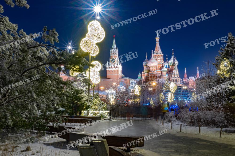 На фотографии изображен зимний вечер в Парке Зарядье, украшенном праздничной иллюминацией. На заднем плане видны башни Кремля и Собор Василия Блаженного. Снежные деревья и светящиеся украшения создают сказочную атмосферу в центре Москвы.