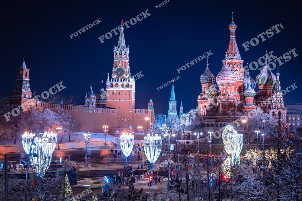 На фотографии изображён Кремль с величественной Спасской башней и Собор Василия Блаженного на Красной площади. Праздничная иллюминация, включающая светящиеся гирлянды и декоративные конструкции, создаёт волшебную атмосферу зимнего вечера. Снежный покров на деревьях и улице усиливает ощущение сказочности. Это место является центром зимних праздников в Москве, привлекая тысячи жителей и гостей города. Традиционная архитектура на фоне ярких огней отражает богатую историю столицы.