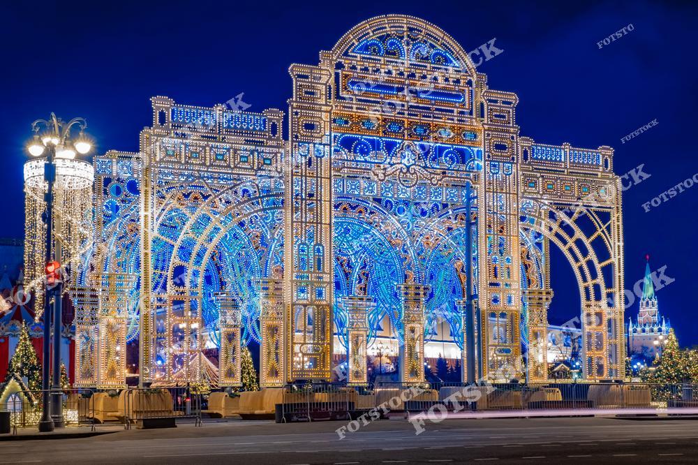 На фото изображена праздничная световая арка в центре Москвы, оформленная в честь Нового года. Эта архитектурная инсталляция, выполненная из тысяч огоньков, создаёт эффект сказочного портала. Вдали виднеются кремлёвские башни, подчёркивая историческую значимость локации. Украшенные ёлки и фонари добавляют уюта и торжественности общей картине. Такой световой декор традиционно устанавливается в период новогодних и рождественских праздников на главных улицах города. Ночная Москва в это время года выглядит особенно нарядно и привлекает множество горожан и туристов.