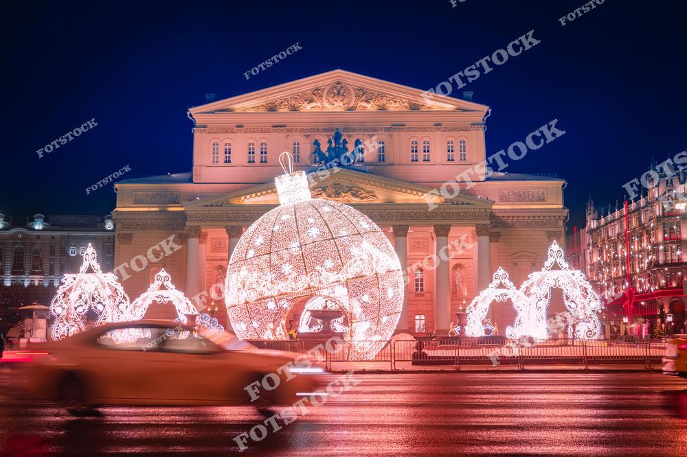 На фотографии изображён Большой театр, одно из самых знаковых зданий Москвы, украшенное новогодними огнями. На переднем плане расположен огромный светящийся шар, через который можно пройти, а также изысканные арки, дополняющие праздничную атмосферу. Театральная площадь преобразилась в зимнюю сказку благодаря яркой иллюминации. Фасад театра подсвечен, подчёркивая его величественную архитектуру. Это место становится центром притяжения для москвичей и туристов в период новогодних праздников.