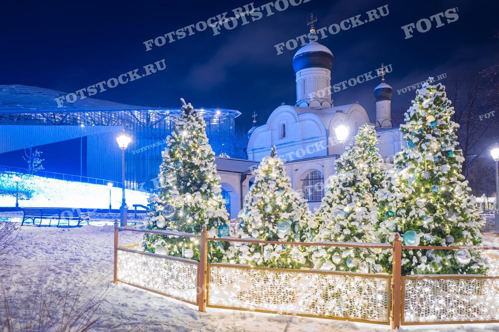 На фотографии изображена зимняя ночь в Парке Зарядье. На переднем плане видны украшенные рождественские елки, покрытые снегом, а на заднем плане — храм, подсвеченный огнями. Праздничная иллюминация и снег создают волшебную атмосферу в центре Москвы.