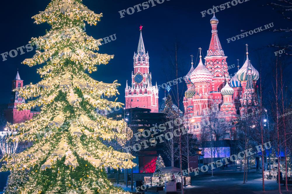 На фотографии запечатлен великолепный вид на московский Кремль. На переднем плане видна пышно украшенная елка, излучающая тёплый свет, который создаёт атмосферу праздника и волшебства. За елкой возвышаются исторические здания: Спасская башня Кремля и собор Василия Блаженного, освещённые разноцветными огнями. Эта фотография идеально подходит для использования в материалах, посвящённых новогодним и рождественским праздникам в России.