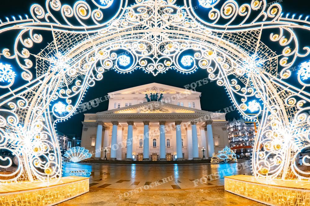 На фотографии изображен Большой театр в Москве, украшенный новогодней иллюминацией. Светящаяся арка на переднем плане создает праздничное настроение. Здание театра подсвечено огнями, подчеркивая его величественную архитектуру в ночное время.